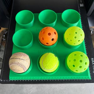 May include: A green plastic jig with nine circular holders, featuring the text "SOFTBALL JIG" and "BASEBALL JIG". The holders contain an orange pickleball, a yellow pickleball, a white baseball, a yellow tennis ball, and a green pickleball.
