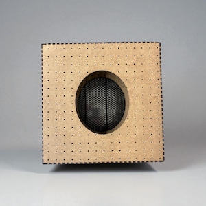 May include: A square, light brown wooden speaker box with a circular black mesh speaker cover. The box has small, evenly spaced holes around the edges and a dark brown border.