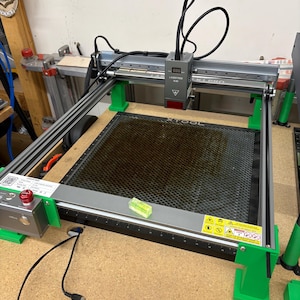 May include: A gray and green laser engraving machine with a black honeycomb work surface. The machine has a digital display, a red power button, and a small level. The machine is on a wooden surface.