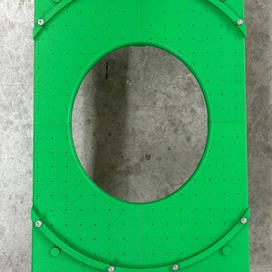 May include: Bright green rectangular plastic object with a large circular cutout in the center. The object has a series of small holes and curved green pieces attached with screws. The object is likely a tool or part of a larger system.