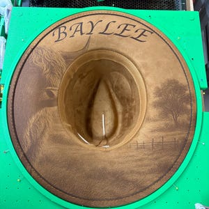 May include: A brown cowboy hat with the name "BAYLEE" engraved on the brim. The hat features a detailed landscape scene with a cow and a tree. The hat is set in a green frame.