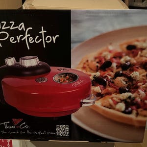 Può includere: Una macchina per pizza rossa e nera con la scritta "Pizza Perfector" sulla scatola. La scatola presenta anche una foto di una pizza cotta. È visibile anche il marchio "Theo & Co.".