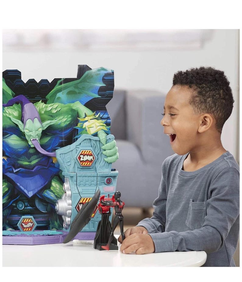 May include: A child is excited while looking at a toy playset featuring a green monster and two action figures. The playset has a blue and gray structure with the text "Zzmp!" and "Twip!" on it. One action figure is black and red.