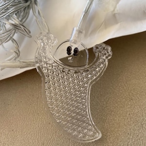 Puede incluir: Luces de cadena de Halloween de plástico transparente con un diseño de fantasma. Cada fantasma tiene una cara sonriente con dos ojos negros. Las luces están ensartadas en un cable plateado.