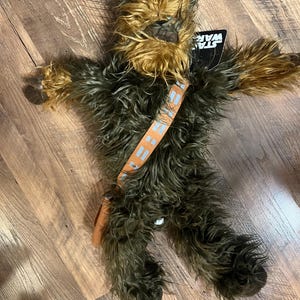 Op de afbeelding: Een Chewbacca pluche pop uit Star Wars. De pop heeft bruin en zwart haar, een beige bandolier en een klein bruin zakje. Er is een Star Wars-label aan bevestigd.