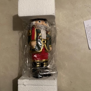 May include: A decorative nutcracker figurine in a plastic bag, packaged in white foam. The nutcracker is dressed in a black and green jacket, red pants, and gold accents. The packaging includes a warning about suffocation.