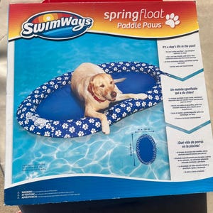 Pode incluir: Um flutuador inflável azul e branco para cães com pegadas de patas. O flutuador é projetado para cães relaxarem na piscina. Tem 104 cm de comprimento e 56 cm de largura. O texto na caixa diz "Swimways spring float Paddle Paws".