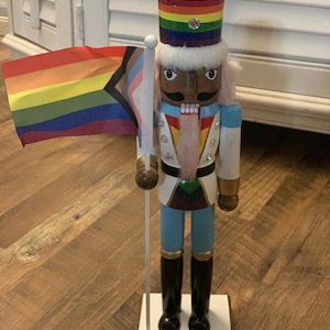 May include: A wooden nutcracker soldier with a rainbow-striped hat and a flag with rainbow and transgender pride colors. The soldier wears a white jacket, blue pants, and black boots. The base of the nutcracker is white and has the year 2022 printed on it.