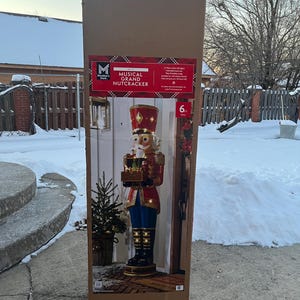 Puede incluir: Una caja de cartón grande que contiene una decoración navideña Musical Grand Cascanueces de 183 cm de altura. El cascanueces viste una chaqueta roja, pantalones azules y botas negras. La caja está sobre una superficie nevada.
