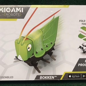 May include: Boxed KAMIGAMI ROBOTS kit featuring a green grasshopper robot named BOKKEN. The box includes the text "FOLD & SNAP TOGETHER!" and "BUILD PROGRAM PLAY." The kit is marked "UNASSEMBLED" and is designed to be programmed via the App Store or Google Play.
