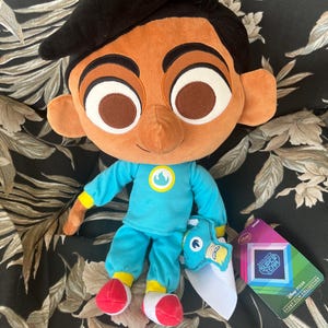 Può includere: Bambola di peluche di un personaggio dei cartoni animati con la pelle marrone, i capelli neri e grandi occhi marroni. La bambola indossa un completo blu con un emblema giallo e scarpe rosse e bianche. È visibile un'etichetta della collezione di cortometraggi "Sanjay's Super Team".