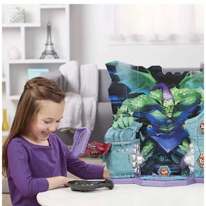 May include: A young girl interacts with a colorful toy playset featuring a large green monster figure. The playset has a purple and blue color scheme with various interactive elements. A black remote control is on the table.