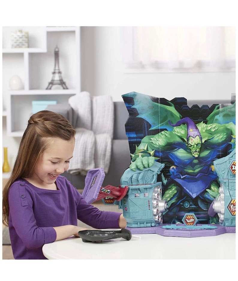 May include: A young girl interacts with a colorful toy playset featuring a large green monster figure. The playset has a purple and blue color scheme with various interactive elements. A black remote control is on the table.