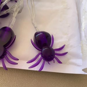 May include: String of purple spider-shaped lights. Each spider has a glossy, dark purple body and lighter purple legs. The lights are strung on a clear wire, perfect for Halloween or spooky decor.
