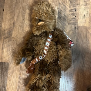 May include: A plush Chewbacca doll, featuring brown fur and a bandolier with white and brown details. The doll has a detailed face and is lying on a wooden surface. A small tag is attached to the doll.