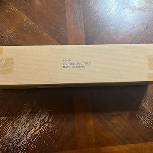 Op de afbeelding: Een rechthoekige kartonnen doos met de tekst "46406 LIGHTED EGG TREE MADE IN CHINA" aan de zijkant. De doos is verzegeld met tape en ligt op een houten oppervlak.