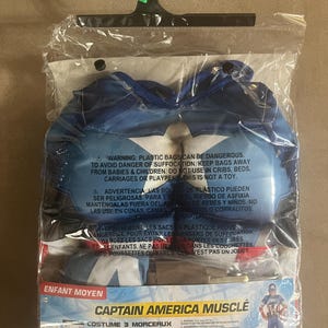 May include: A Captain America muscle costume for children, packaged in a clear plastic bag. The costume includes a mask, belt, and muscle suit. The packaging features the Marvel logo and a graphic of Captain America.