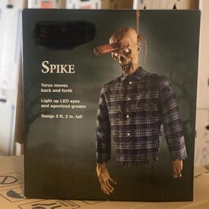 May include: A black and white box featuring a hanging animated prop. The prop is a man wearing a plaid shirt with glowing eyes. The box reads "SPIKE" and describes the prop as having a moving torso, light up LED eyes, and agonized groans. The prop is 3 feet 2 inches tall.