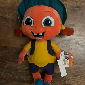 May include: A plush doll with an orange face, big eyes, and a smiling mouth. It wears a yellow shirt, blue shorts, and a teal hat. The doll has a tag attached.