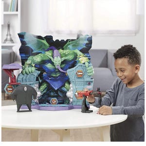 May include: A child plays with a toy playset featuring a large, green monster figure with a purple hat and tongue. The playset includes a figure of a man in a black suit and a red and black toy weapon. The background features a cityscape.