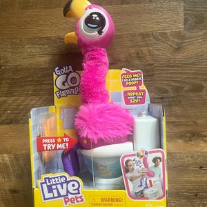 May include: A pink and yellow Gotta Go Flamingo toy with a toilet playset. The toy has a fluffy pink neck and head, with large eyes and a yellow beak. The packaging includes the text "Little Live Pets" and "Feed Me! I Do a magic POOP!"