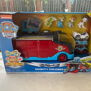May include: A Paw Patrol Mighty Cruiser toy set in a box. The toy features a red and black cruiser with blue accents, a small white vehicle, and several Paw Patrol character figures. The box has the Nickelodeon logo and the text "Mighty Super Paws".