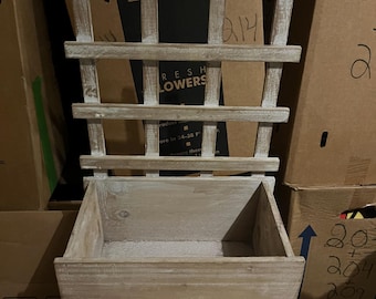 Op de afbeelding: Een verweerde, lichtgekleurde houten plantenbak met een latwerk. De plantenbak is rechthoekig en staat op kleine voetjes. Het latwerk heeft een rasterpatroon. De woorden "FRESH FLOWERS" zijn gedeeltelijk zichtbaar.