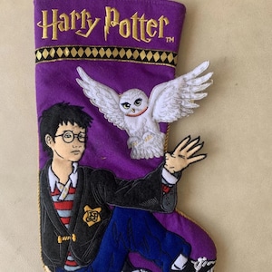 Puede incluir: Calcetín navideño de fieltro morado con Harry Potter y Hedwig. El calcetín tiene adornos dorados y un lazo dorado para colgar. Harry lleva gafas, una chaqueta negra y pantalones azules. El texto "Harry Potter" está bordado en dorado.
