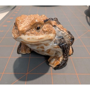 May include: A ceramic toad figurine in shades of brown, tan, and black. The toad has a textured skin appearance and detailed facial features. The figurine is a decorative item, likely for home decor or a collector's piece.