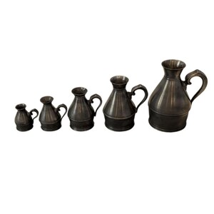May include: A set of five antique metal pitchers in varying sizes. Each pitcher has a dark, metallic finish, a rounded body, a handle, and a flared spout. The pitchers are arranged in a row, showcasing their graduated sizes.