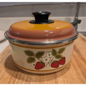 Puede incluir: Una olla de cocina vintage con tapa. La olla tiene una base de color crema con un diseño de fresas y flores. La tapa es naranja y marrón, y el asa es negra. La olla es de metal y tiene un borde plateado.