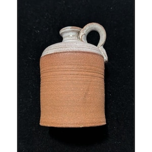 May include: A small, handmade ceramic jug with a handle. The jug has a cylindrical body in a reddish-brown hue, with horizontal textured lines. The top and handle are a light beige color. The jug is likely used for storing liquids.