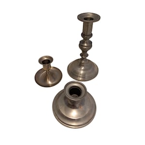 Puede incluir: Tres candelabros plateados de diferentes alturas y diseños están dispuestos sobre una superficie blanca. El candelabro más alto tiene un tallo delgado y adornado. Los otros dos tienen bases redondas más simples. Los candelabros son de metal.