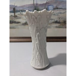 May include: A white ceramic vase with a flared top and a detailed, embossed leaf pattern. The vase has a narrow base and a tall, slender body. The background features a framed landscape painting.