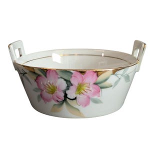 Puede incluir: Cuenco de porcelana blanca con dos asas, con flores rosas pintadas a mano con centros amarillos y hojas verdes. El borde está adornado con oro. Un plato decorativo.