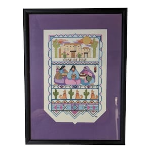 May include: Framed cross-stitch artwork featuring a Southwestern desert scene with cacti, buildings, and figures. The artwork is in a black frame with a purple mat. The text "CASA DE PAZ" is visible in the design.