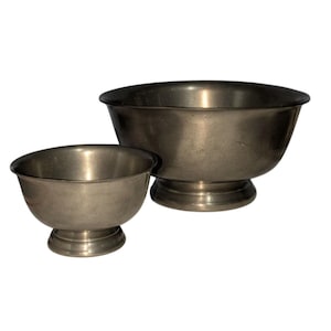 May include: Two antique metal bowls. The bowls are silver-coloured with a slightly tarnished appearance. The larger bowl has a wider rim and sits on a pedestal base. The smaller bowl is similar in design, with a rounded shape and a pedestal base. Both bowls are empty.