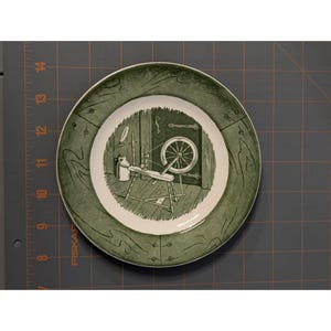 Può includere: Un piatto decorativo rotondo con un design verde e bianco. Il piatto presenta un'illustrazione centrale di un filatoio e altri strumenti, circondata da un bordo verde con un motivo a venature del legno. Il piatto è probabilmente in ceramica.