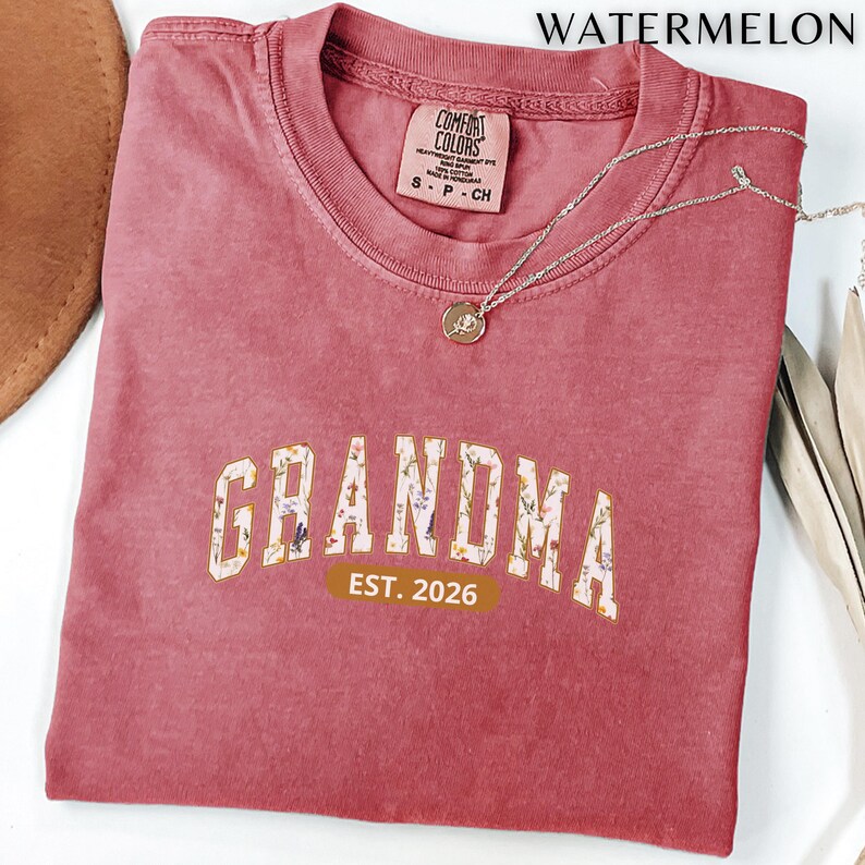 Floral Grandma Est. 2026 shirt with large varsity-style lettering filled with colorful wildflowers, a warm golden outline, and a rounded brown accent beneath the design on a black background.