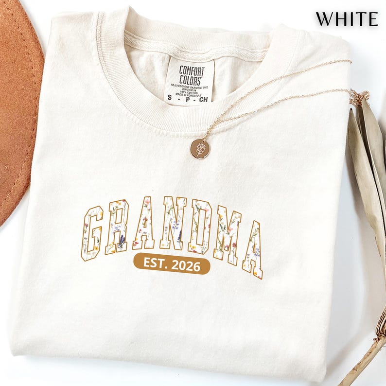 Floral Grandma Est. 2026 shirt with large varsity-style lettering filled with colorful wildflowers, a warm golden outline, and a rounded brown accent beneath the design on a black background.