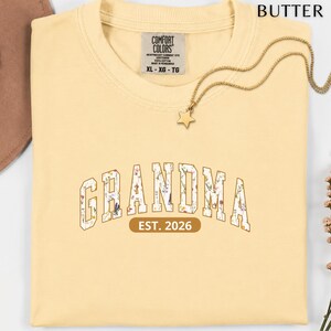 Floral Grandma Est. 2026 shirt with large varsity-style lettering filled with colorful wildflowers, a warm golden outline, and a rounded brown accent beneath the design on a black background.