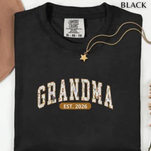 Floral Grandma Est. 2026 shirt with large varsity-style lettering filled with colorful wildflowers, a warm golden outline, and a rounded brown accent beneath the design on a black background.