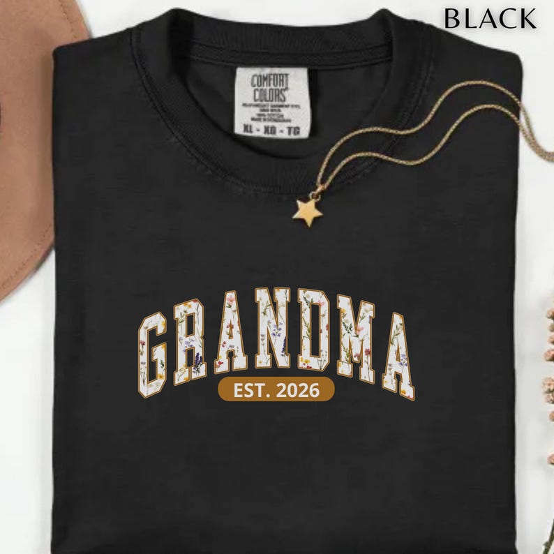 Floral Grandma Est. 2026 shirt with large varsity-style lettering filled with colorful wildflowers, a warm golden outline, and a rounded brown accent beneath the design on a black background.