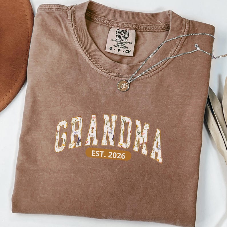 Floral Grandma Est. 2026 shirt with large varsity-style lettering filled with colorful wildflowers, a warm golden outline, and a rounded brown accent beneath the design on a black background.