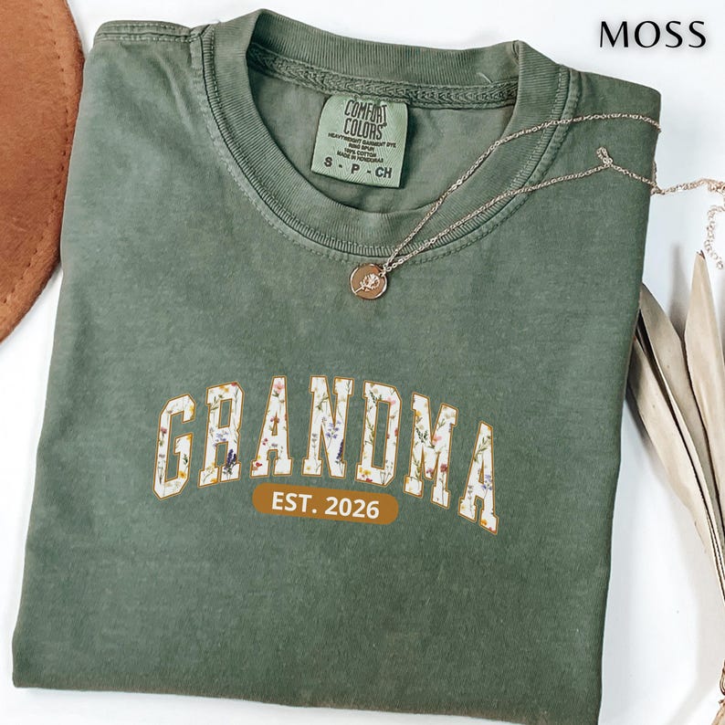 Floral Grandma Est. 2026 shirt with large varsity-style lettering filled with colorful wildflowers, a warm golden outline, and a rounded brown accent beneath the design on a black background.