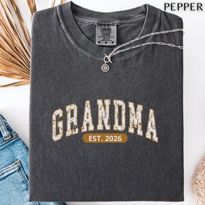 Floral Grandma Est. 2026 shirt with large varsity-style lettering filled with colorful wildflowers, a warm golden outline, and a rounded brown accent beneath the design on a black background.