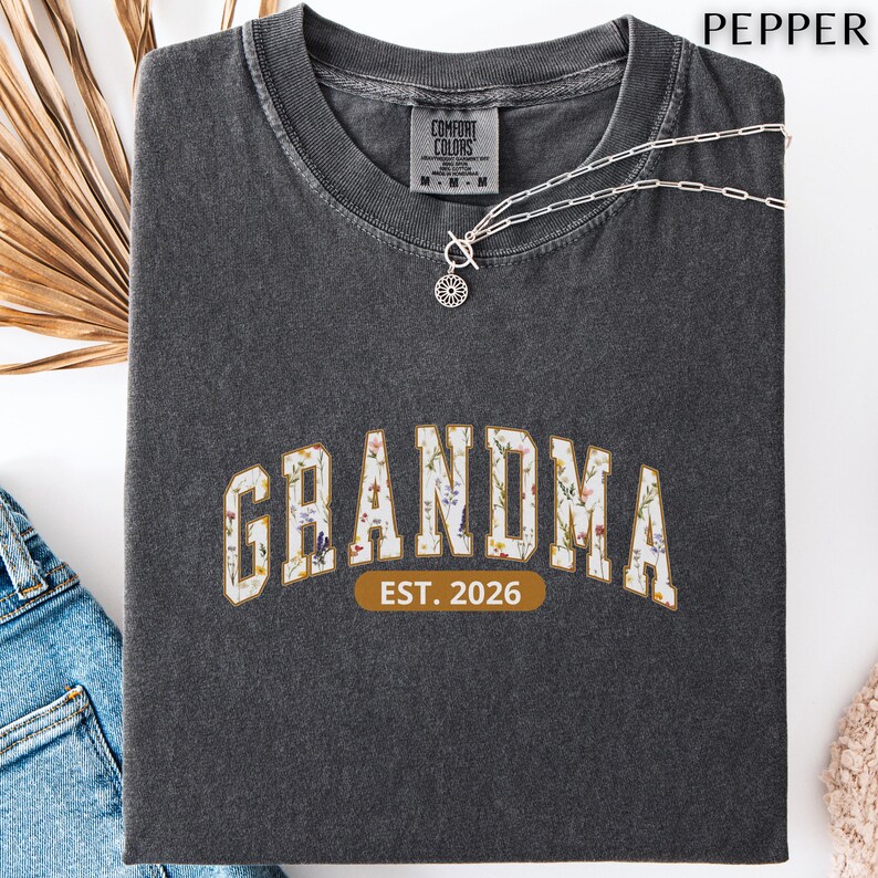 Floral Grandma Est. 2026 shirt with large varsity-style lettering filled with colorful wildflowers, a warm golden outline, and a rounded brown accent beneath the design on a black background.