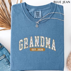 Floral Grandma Est. 2026 shirt with large varsity-style lettering filled with colorful wildflowers, a warm golden outline, and a rounded brown accent beneath the design on a black background.