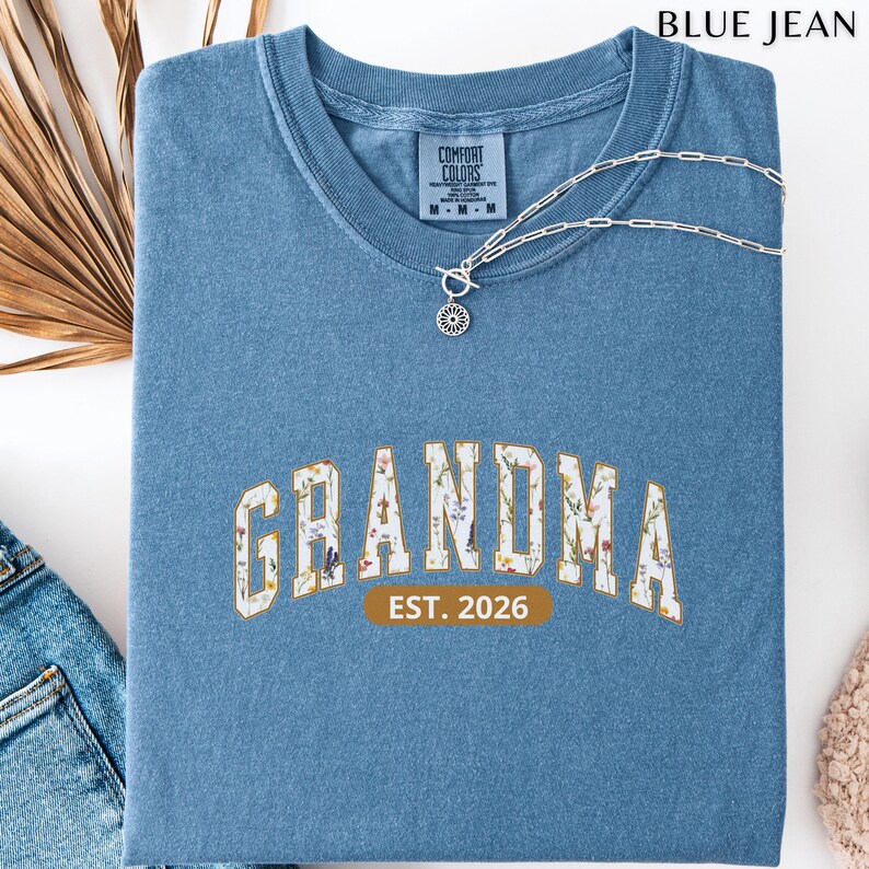 Floral Grandma Est. 2026 shirt with large varsity-style lettering filled with colorful wildflowers, a warm golden outline, and a rounded brown accent beneath the design on a black background.
