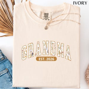 Floral Grandma Est. 2026 shirt with large varsity-style lettering filled with colorful wildflowers, a warm golden outline, and a rounded brown accent beneath the design on a black background.